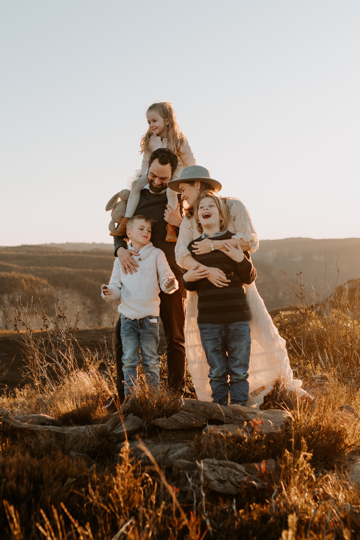 Family of five sharing a joyful moment on a mountaintop at sunset in the Blue Mountains, NSW, with golden light and scenic views in the background.