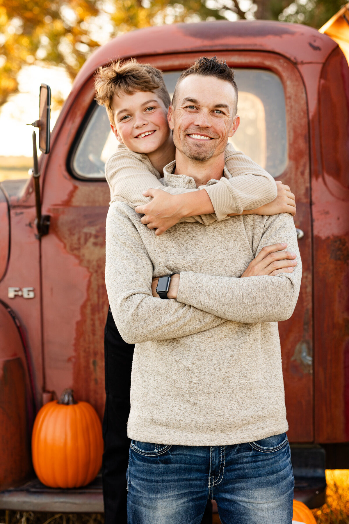 Preteen boy stands behind his dad and hugs his dad around the neck as they both smile at the camera.