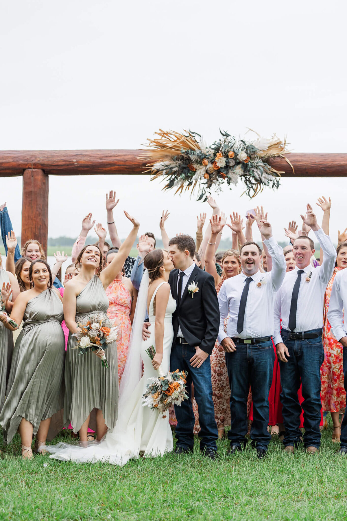 Bride and groom exchange a kiss while their friends and family gather around them, cheering and waving their arms in the air.