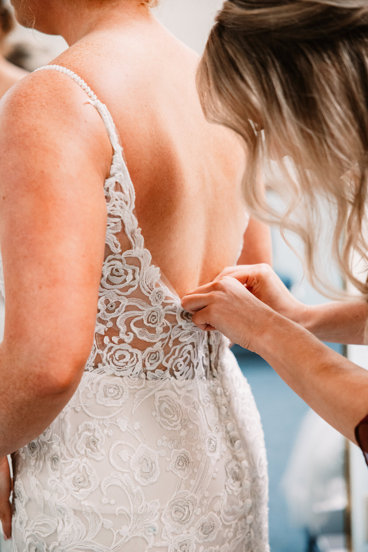 A bride's lace gown is buttoned while she stands