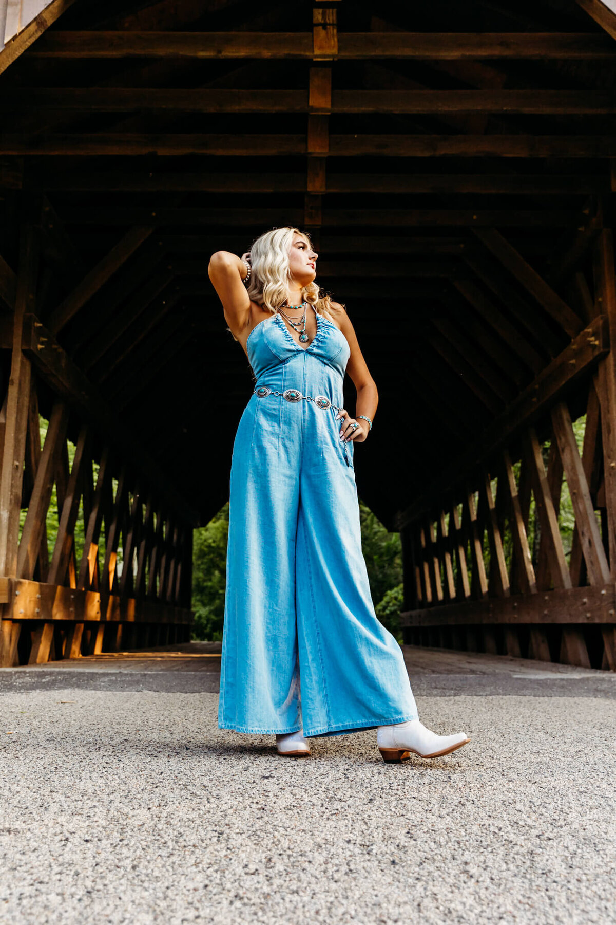 stunning teenage girl in a blue jean jumpsuit and white cowboy boots standing in a power pose as she plays with her hair and looks off into the distance for her senior photography session with Ashley Kalbus.