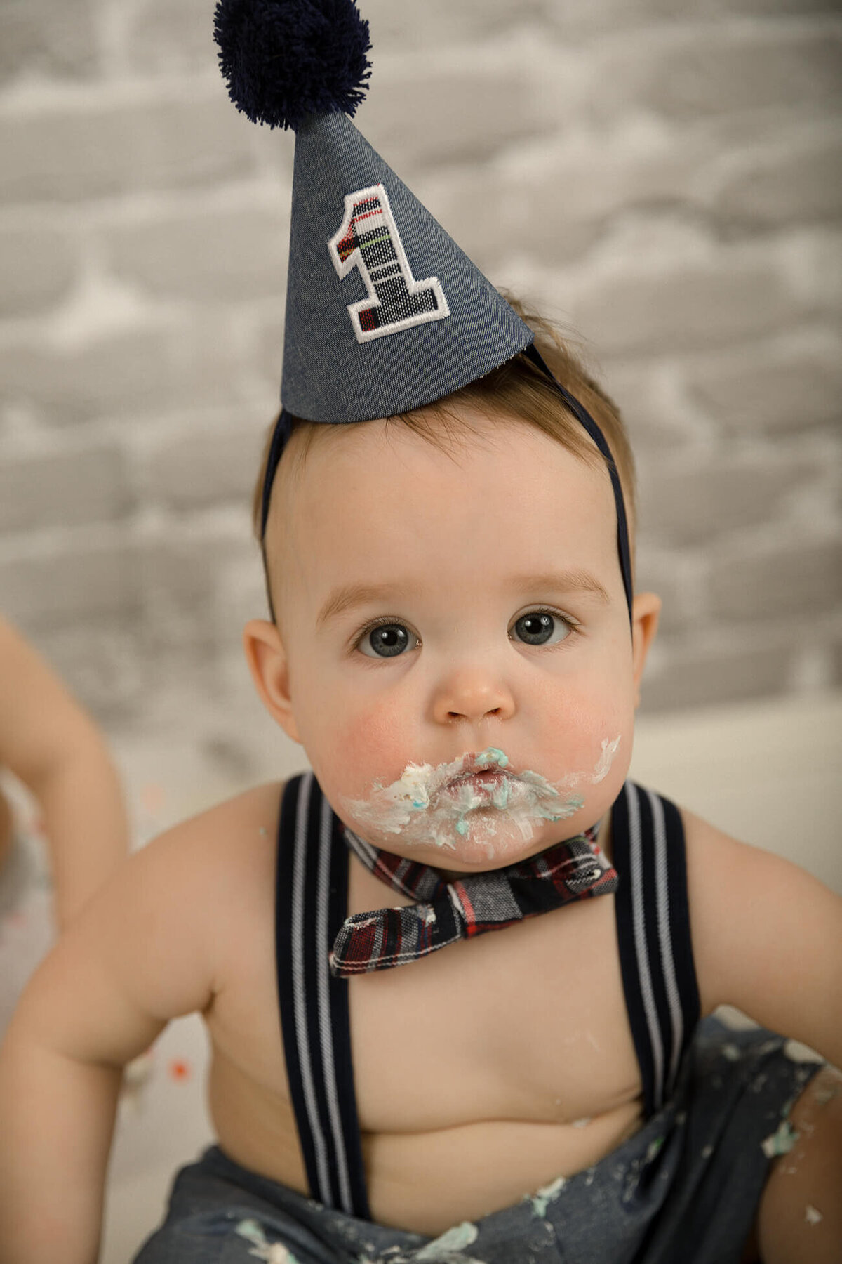 1 year old boy with blue suspenders and party hat with lots of frosting on his face.