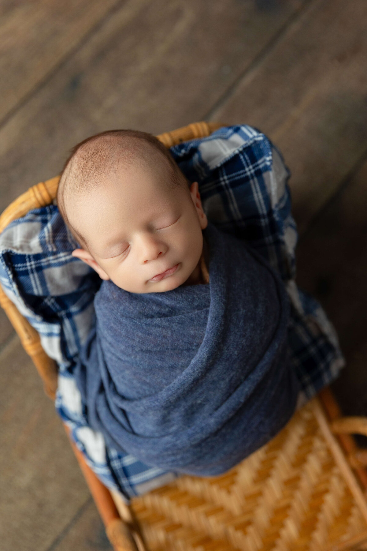A newborn baby is nestled in a woven basket lined with a blue plaid blanket. The baby, wrapped in a dark blue blanket, has closed eyes and a serene expression, creating a tranquil atmosphere.