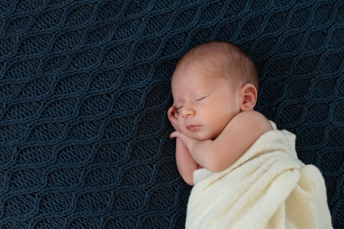 A newborn baby sleeps on its side on a blue knit blanket