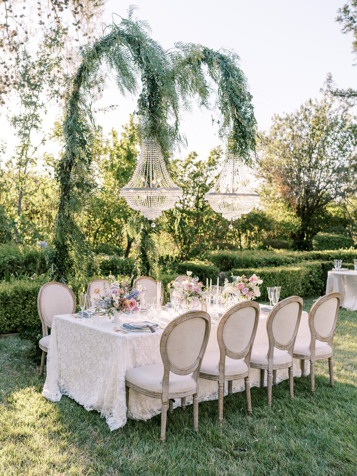 A table set for a gorgeous whimsical Bridgerton inspired wedding at Chateau Robles's in Paso Robles, California with overhanging chandeliers, covered with vines and exquisite lace from BBJ