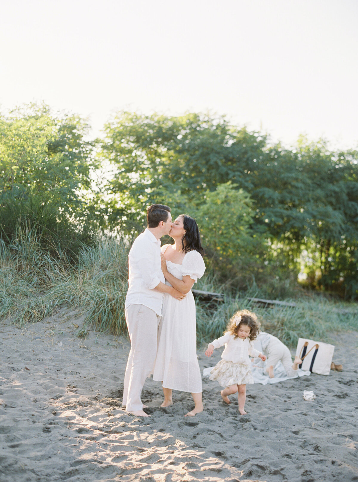 mom and dad kissing on the beach while toddlers run around them at this Family photo session in Seattle.