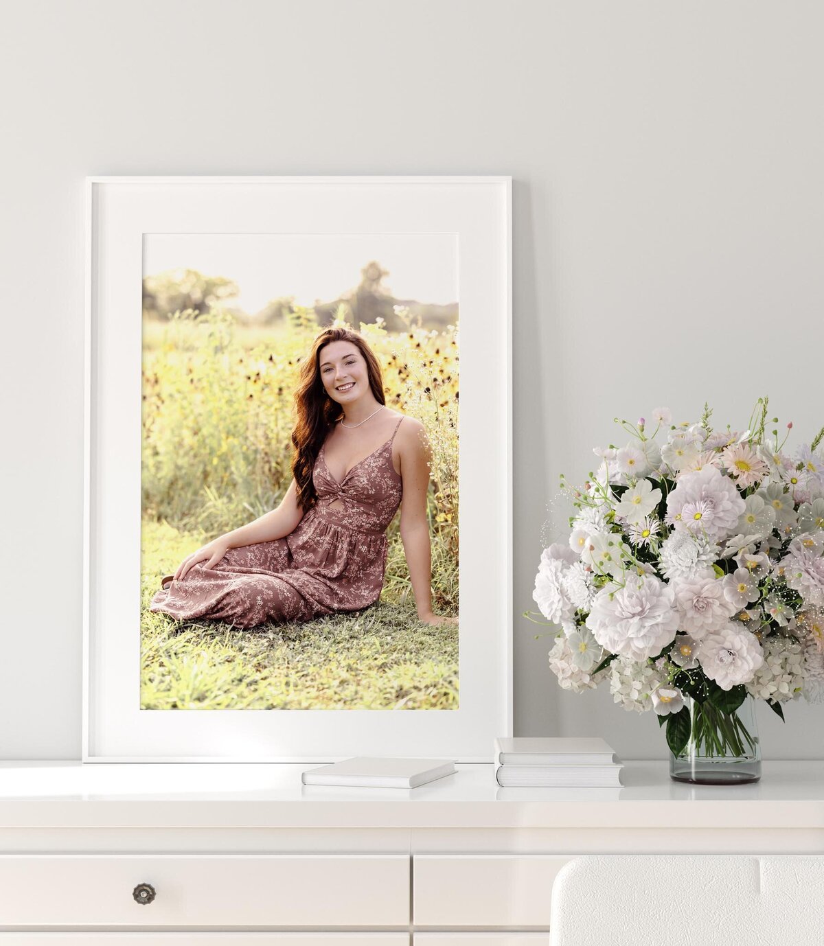 A portrait of a high school senior in a flower field. The framed picture sits on a white desk with a vase of white flowers next to it.