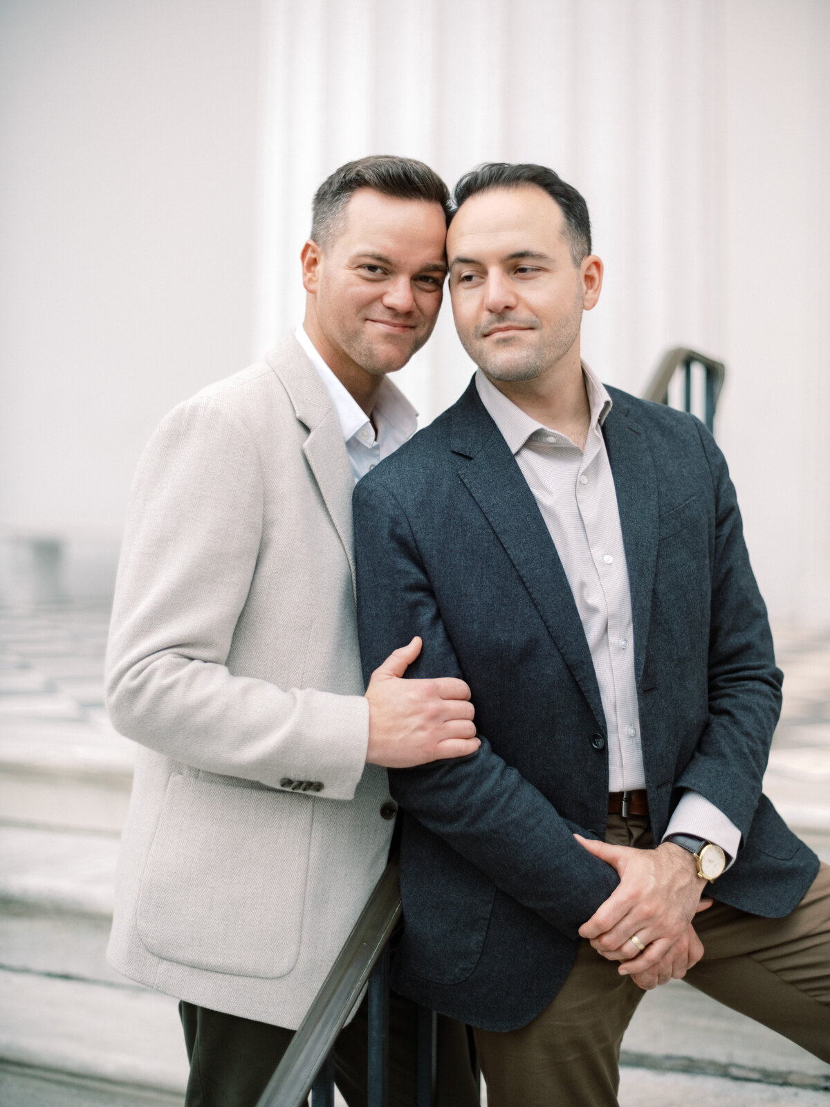 A stylish same-sex couple stands close, embracing each other in an elegant setting, showcasing their love and connection against a stunning architectural backdrop that enhances their aura.