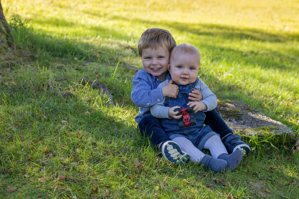 Søskenfoto av to brødre som sitter på gresset og ser mot kamera. Lillebror på et år sitter på fanget til storebror på tre.