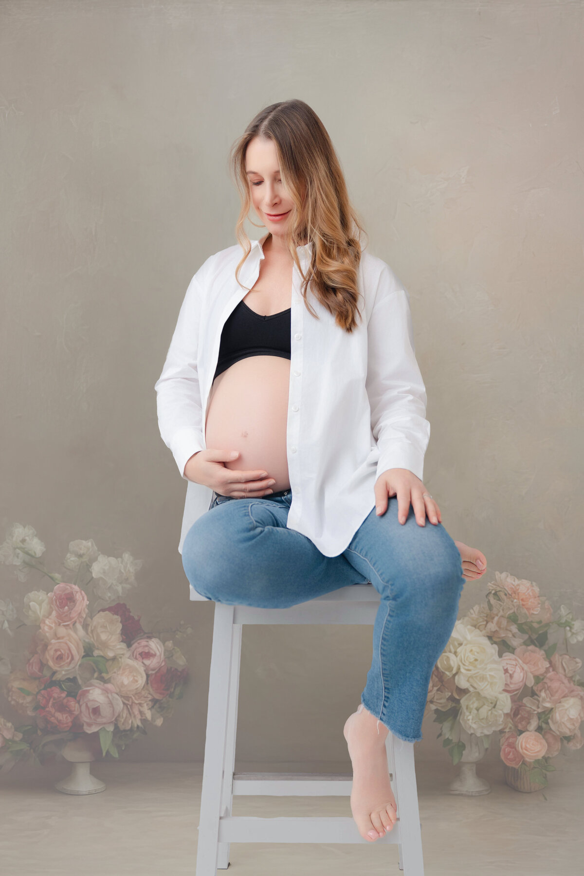 Pregnant posed mom sitting on a stool and wearing a white oversized shirt unbuttoned and showing black sports bra.