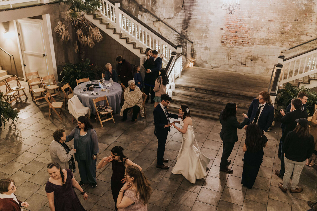 A bride and groom dance in the center of a warmly lit courtyard, surrounded by guests who are talking, sitting at tables, or dancing. The space features stairs, greenery, and exposed brick walls.