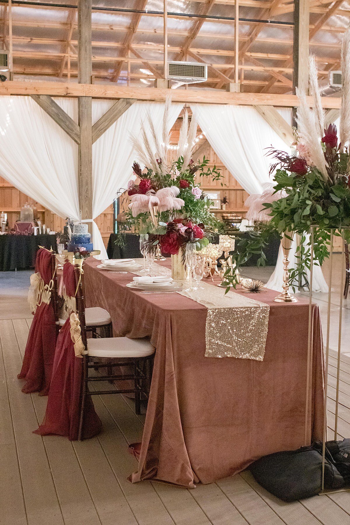 The sweetheart table, set on the stage at Saddle Woods Farm is set with a blush velvet table cloth and a gold sequined table runner. The brown chiavari chairs are set with white cushions and are decorated with burgundy organza fabric and gold BRIDE and GROOM signs hanging on the back. The table has two large blush and burgundy floral arrangements on gold stands accented by pampas grass.