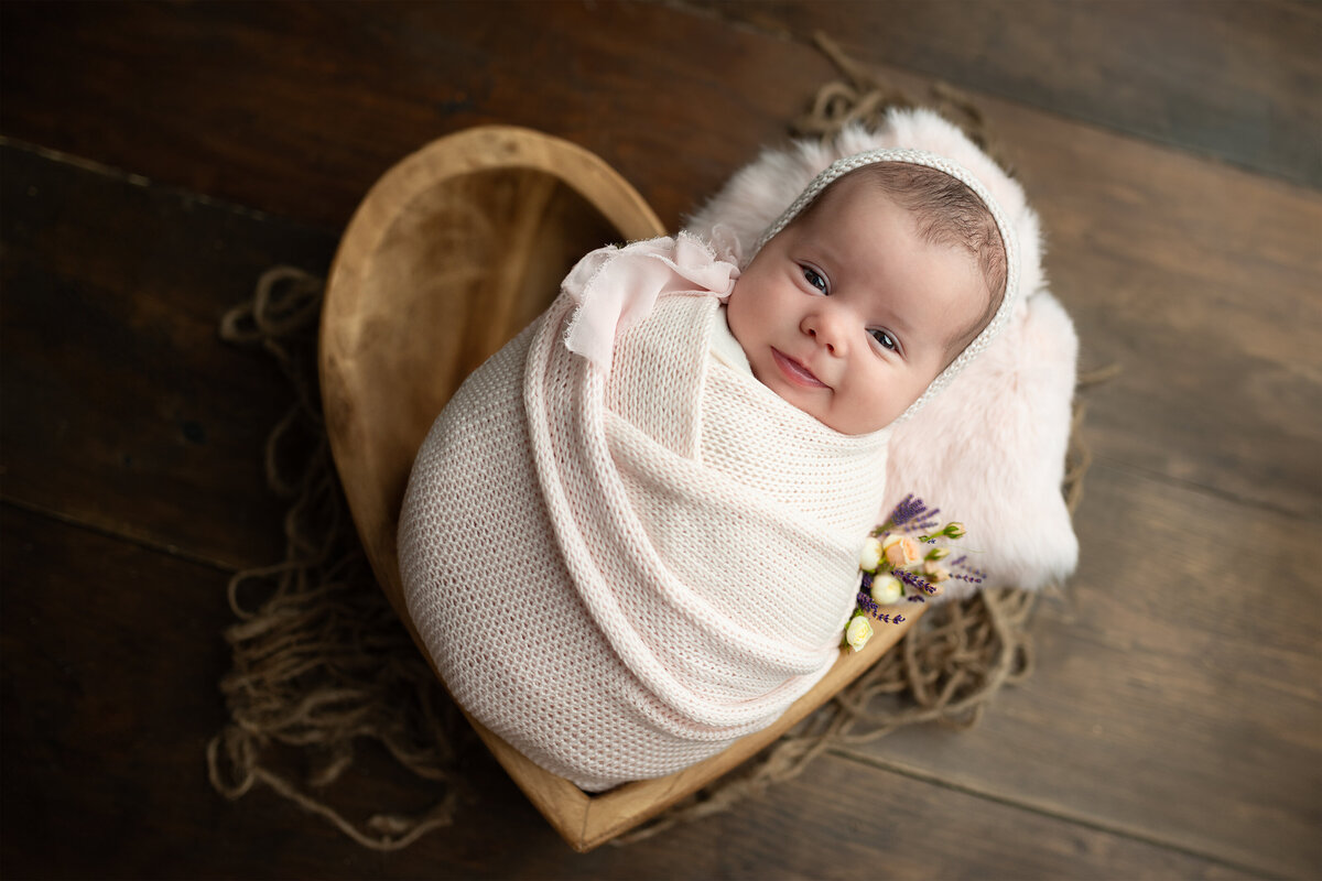 Heartfelt client testimonial for newborn photography featuring a swaddled baby in a pink wrap, nestled in a handcrafted wooden heart bowl with floral details. Captured in a warm, rustic-styled photography session at Meraki Photographic Creations Studio in Paris, Ontario.