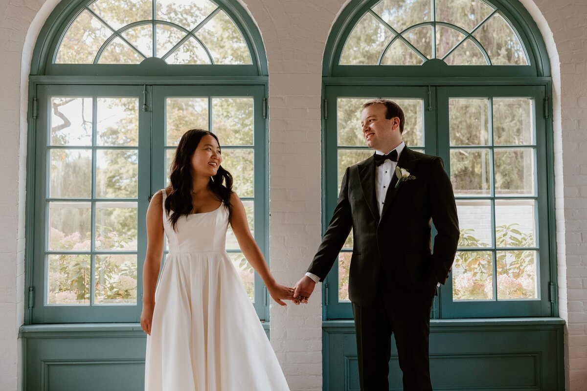 bride and groom stand hand in hand in front of large arched windows at Stagecoach Inn. The bride wears a simple, elegant gown while the groom is dressed in a classic black tuxedo. They share a joyful glance with each other, framed by the natural light coming through the windows and the greenery outside. The bright, airy space adds a romantic and timeless feel to the moment.