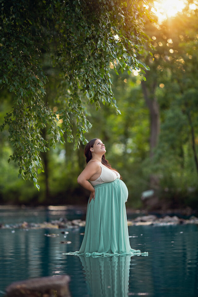 An expecting mother stands in a pool of water under a tree at sunset in a blue maternity dress