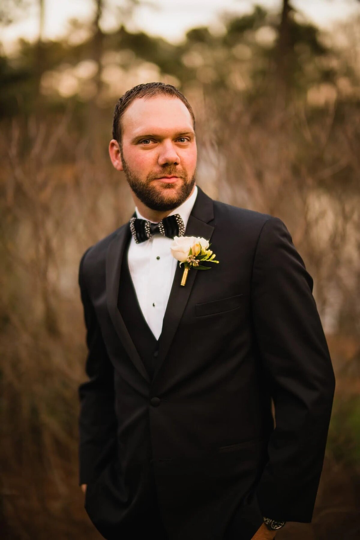 A groom in a black tuxedo with a bow tie stands confidently outdoors