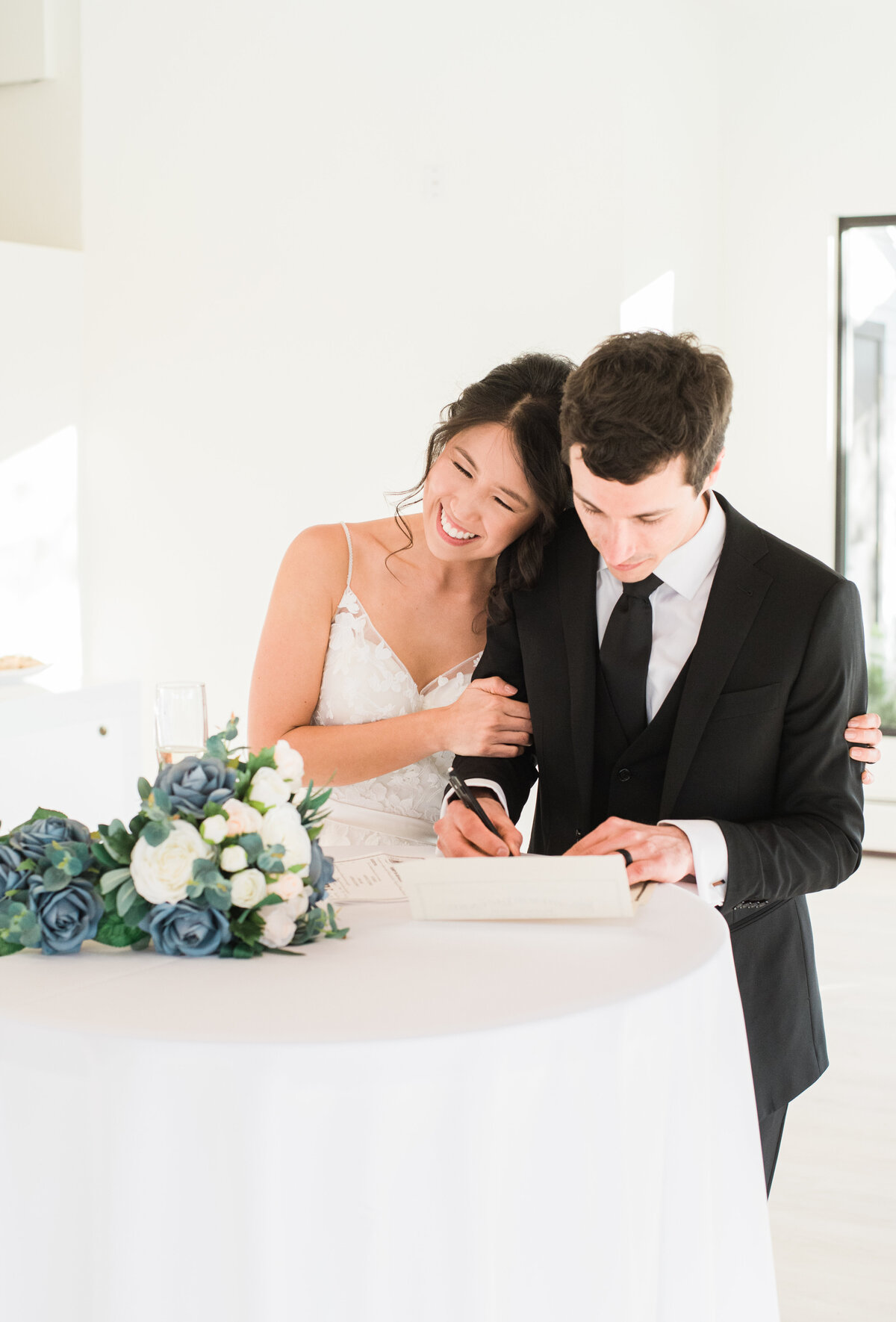 At The Oaks, captured by Denver wedding photographer KB Radiant photography, a bride and groom sign their marriage license while the bride rests her head on the groom as he signs