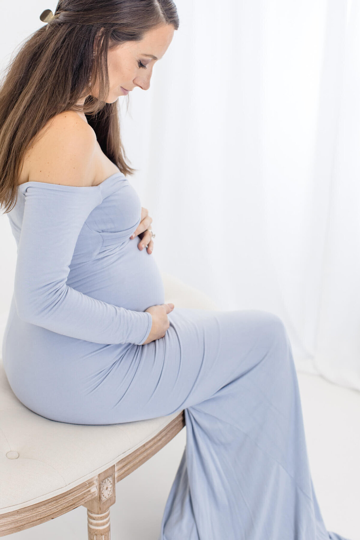 A pregnant woman in a light blue, off-the-shoulder dress sits on a cushioned bench, gently holding her baby bump. She has long brown hair and gazes down with a serene expression. The background is softly lit and white.