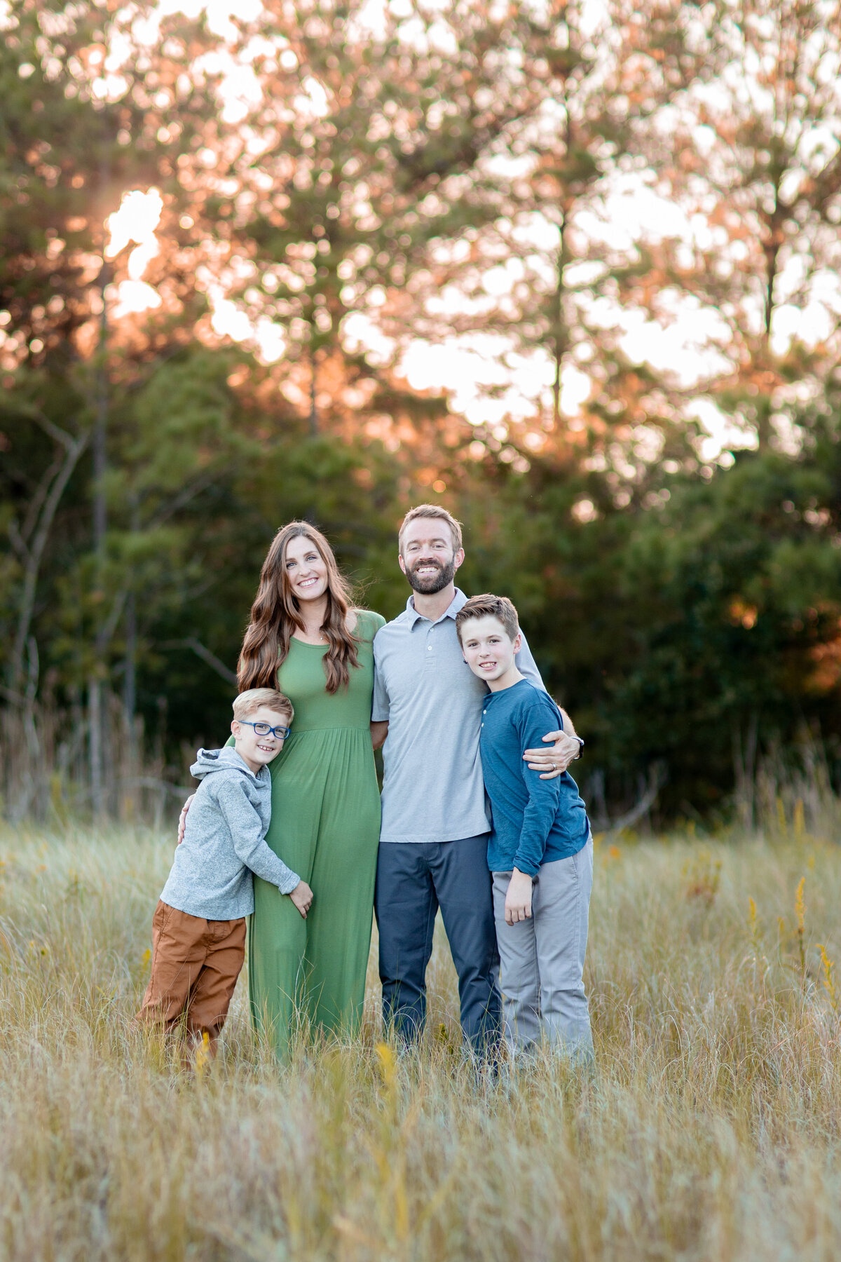 A joyful family of four stands close together in a golden field during fall in Virginia, dressed in coordinated seasonal outfits as the sun sets behind them- capturing connection, color, and the love that binds them. 