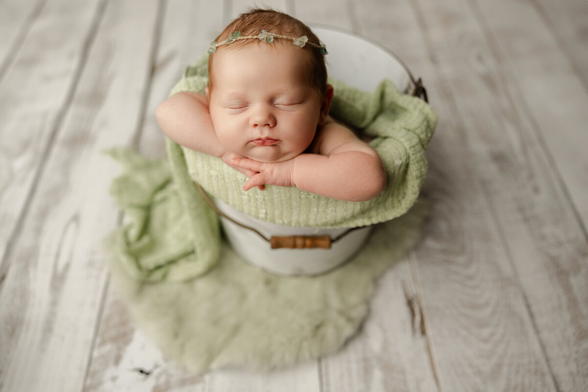 Newborn baby girl resting peacefully in a white bucket, styled with sage green knits and a dainty floral headband. Captured by Inland Empire newborn photographer Kaitlyn Dawn Photography.