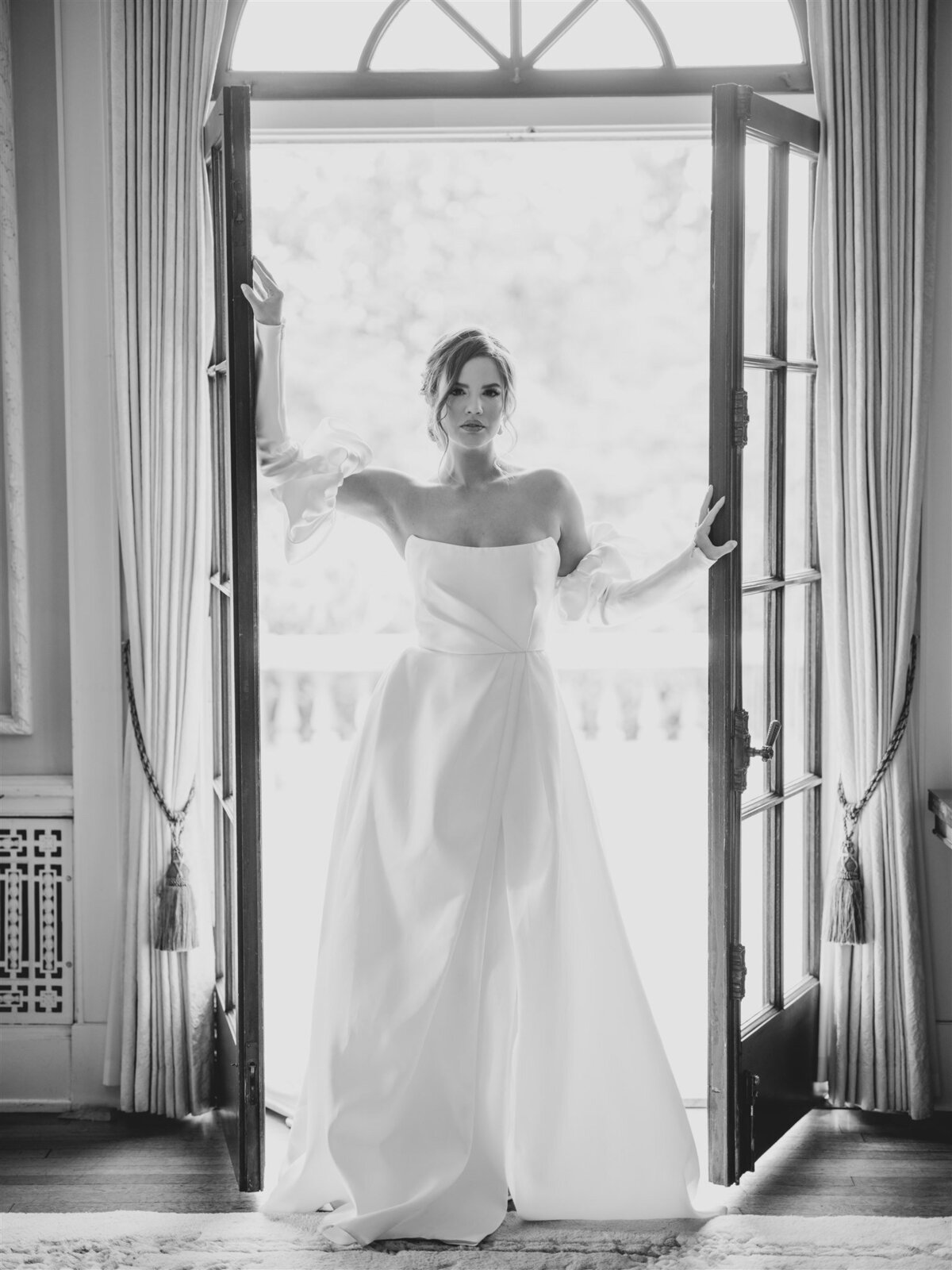 A woman stands in a doorway, wearing an elegant, off-the-shoulder white gown with long, sheer sleeves. Captured in black and white, the moment reflects the timeless style of destination wedding planner Melissa Dawn of Event Designs.