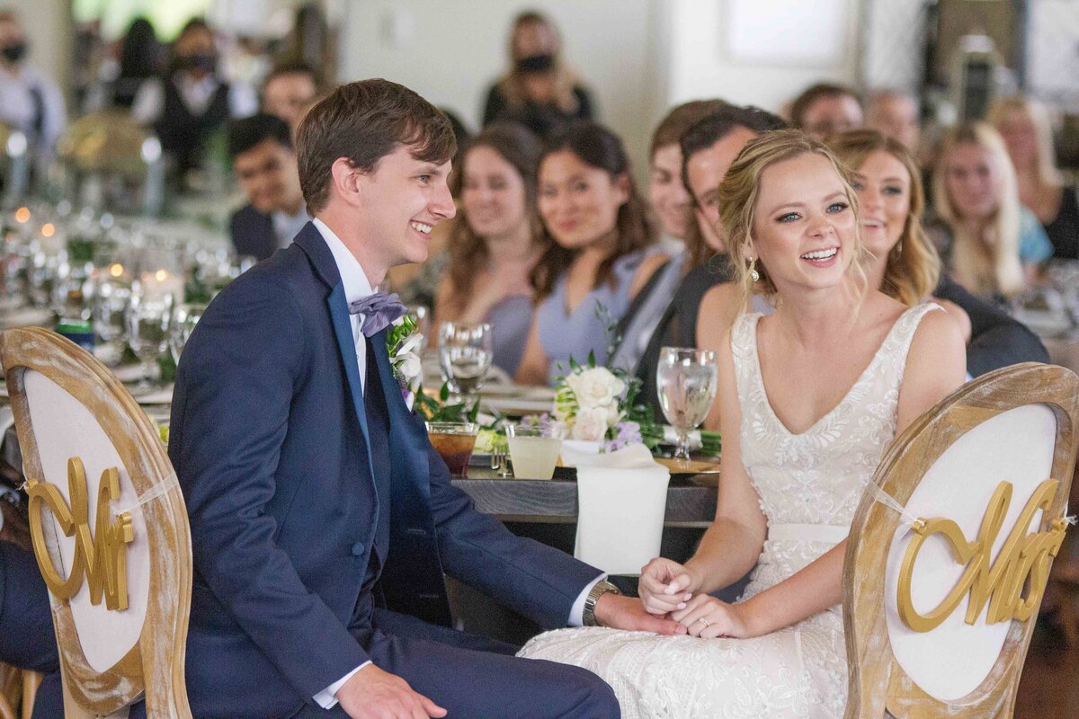 bride and groom hold hands listening to speech in oval backed chairs  by wedding photographer in Texas