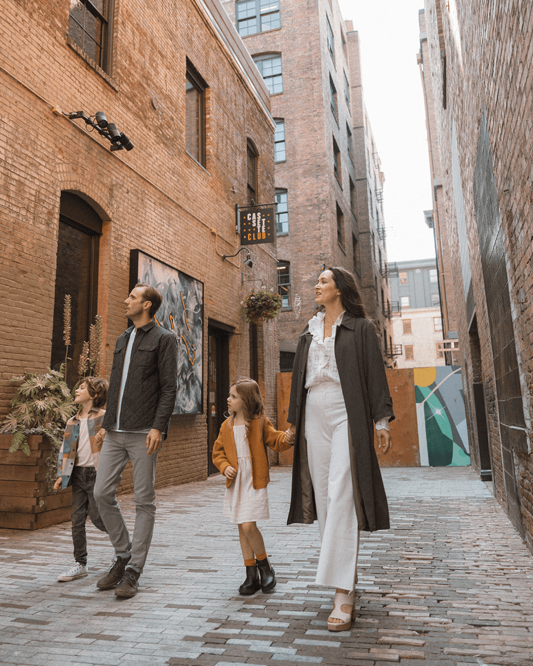 Short Story mini session in Orange County — candid family photo of parents and children walking together in a city alley with warm fall outfits.