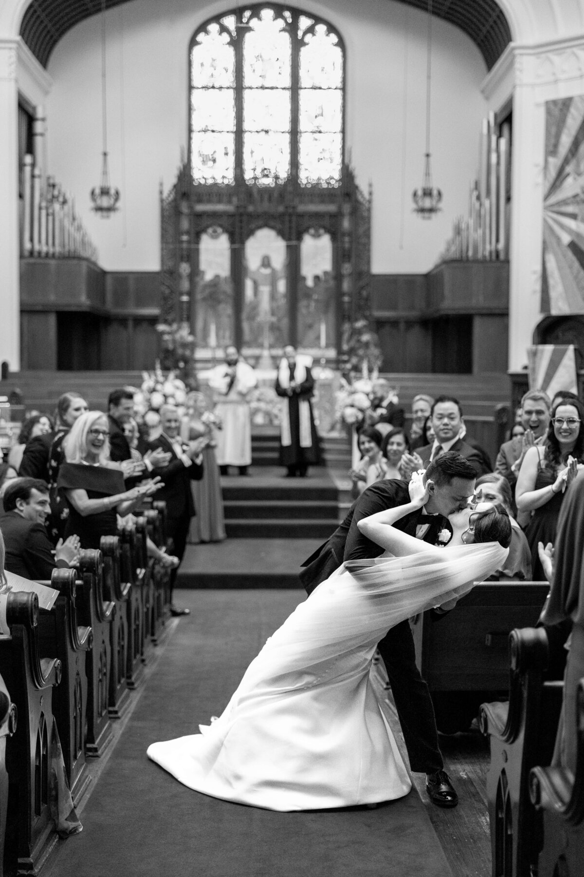 A couple shares a romantic kiss in the aisle of a church during their wedding ceremony. Guests seated in pews smile and clap. The altar and stained glass window are visible in the background. The scene is in black and white.