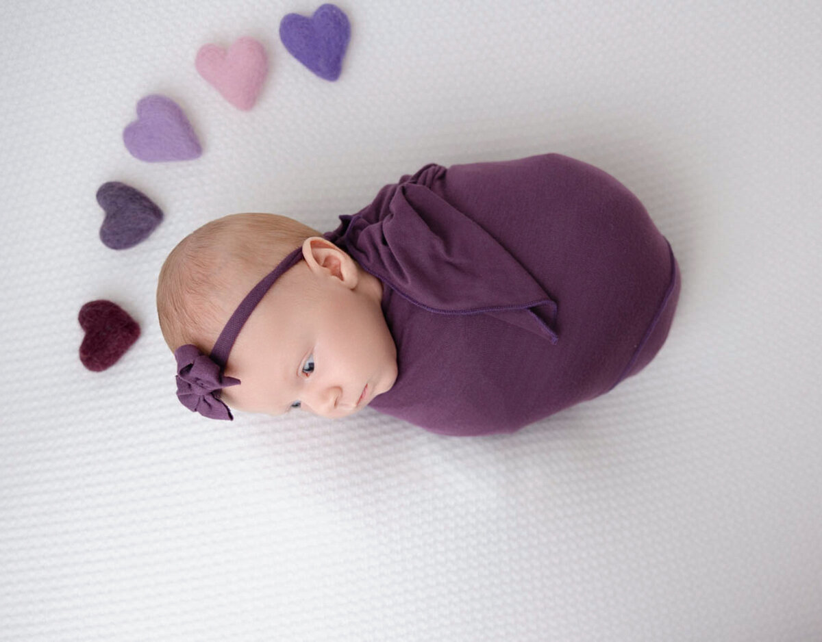 two-week baby girl posed lying on white blanket and wearing a purple head bow and wrap.  Behind baby are five different colored hearts behind baby.