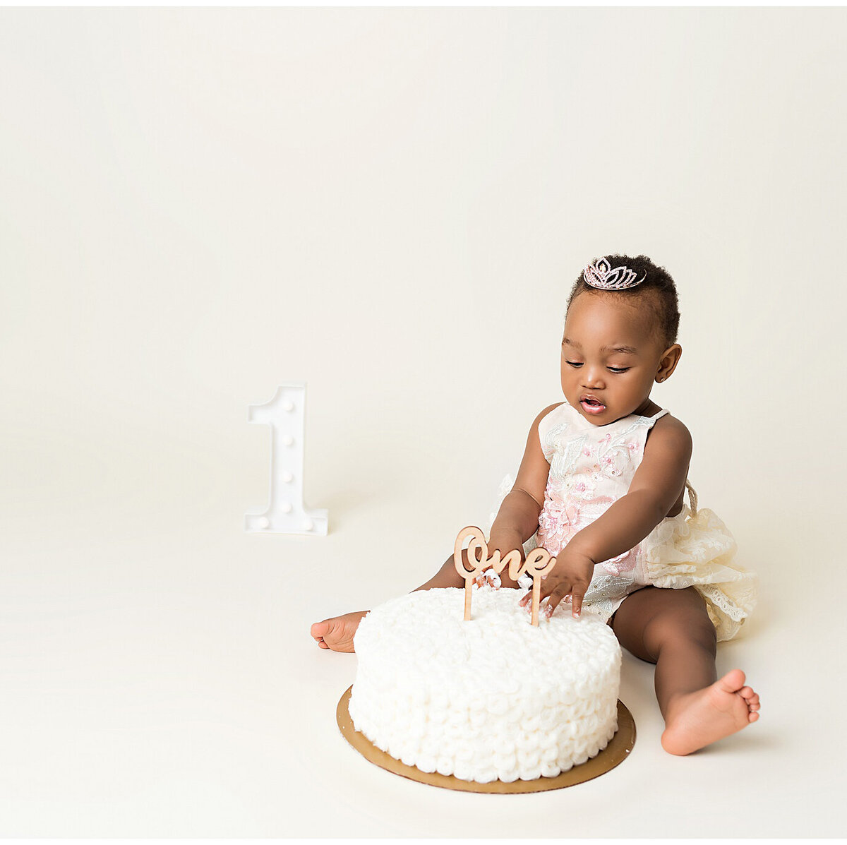 A little girl sitting on the floor with hands on cake. A little number one in the back.