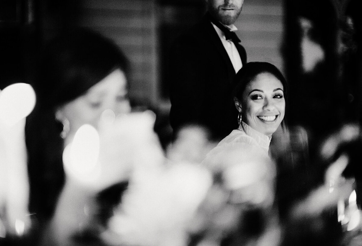 A smiling woman, a man in the background, and a blurred image of another woman in front of her