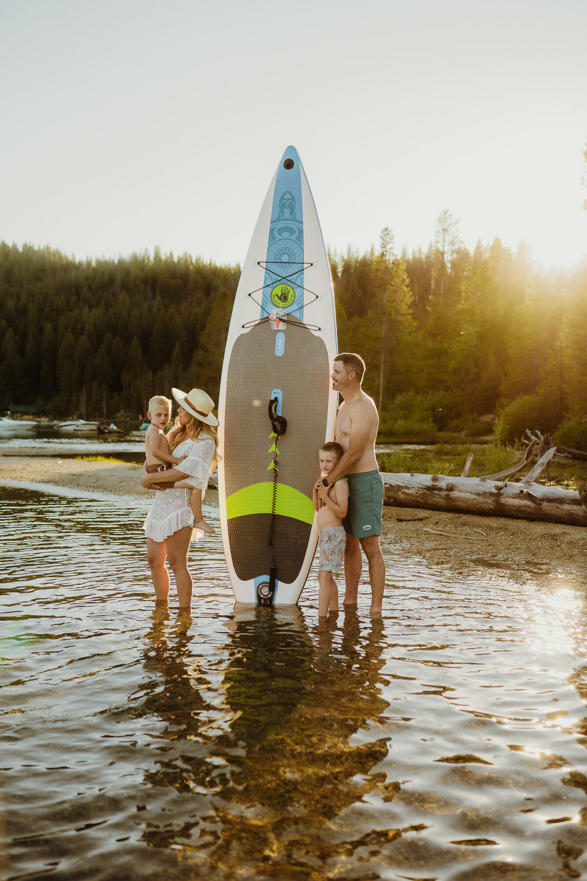 A family of four is standing around their paddle board in the water as the sun dips behind the mountains