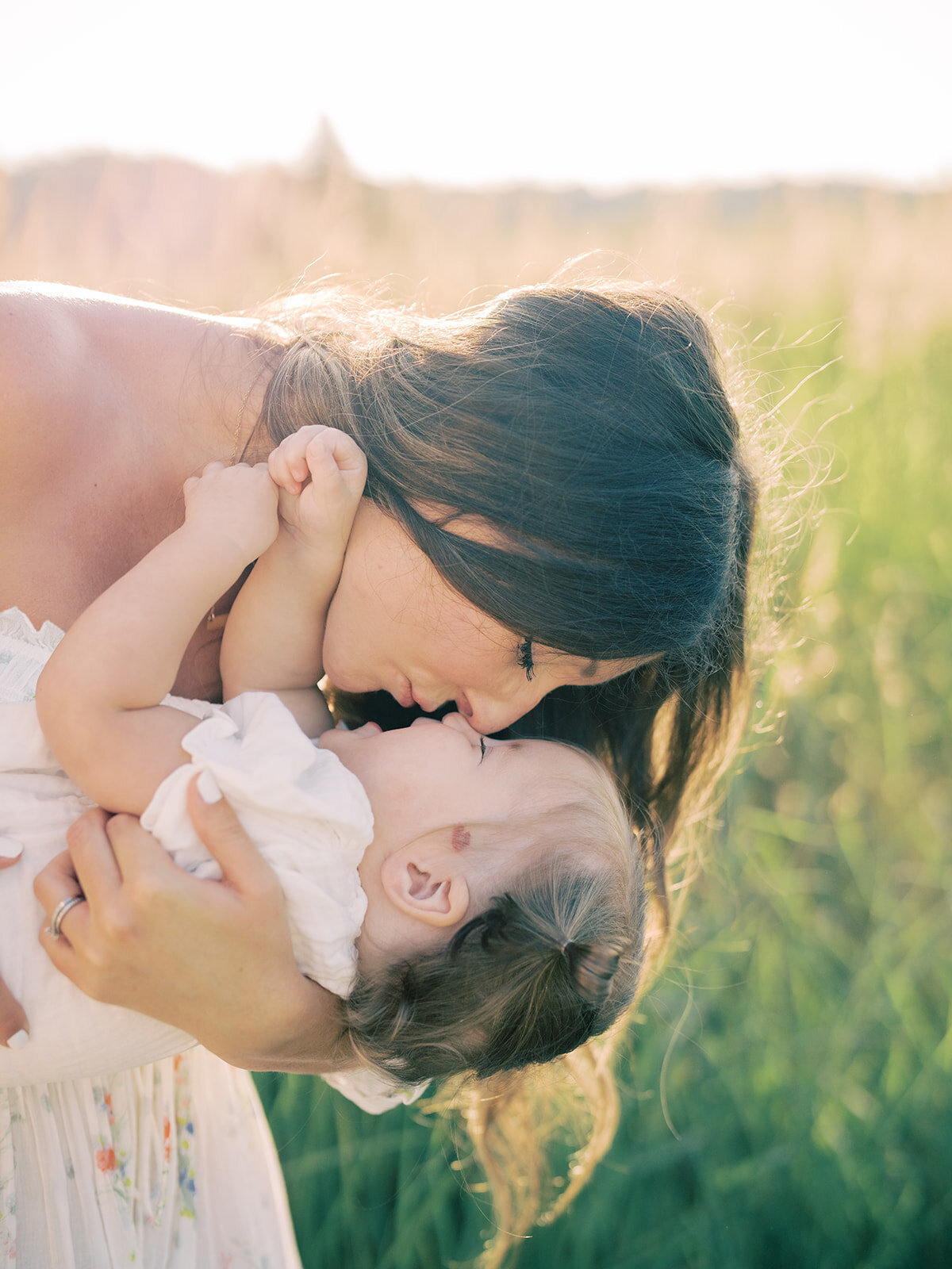 Mother leans down holding her toddler daughter in a grassy field at sunrise.