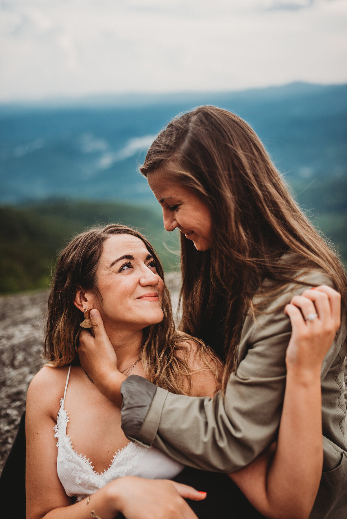 a LGBTQIA+ couple sitting in an embrace on a mountain with a mountain range in the background