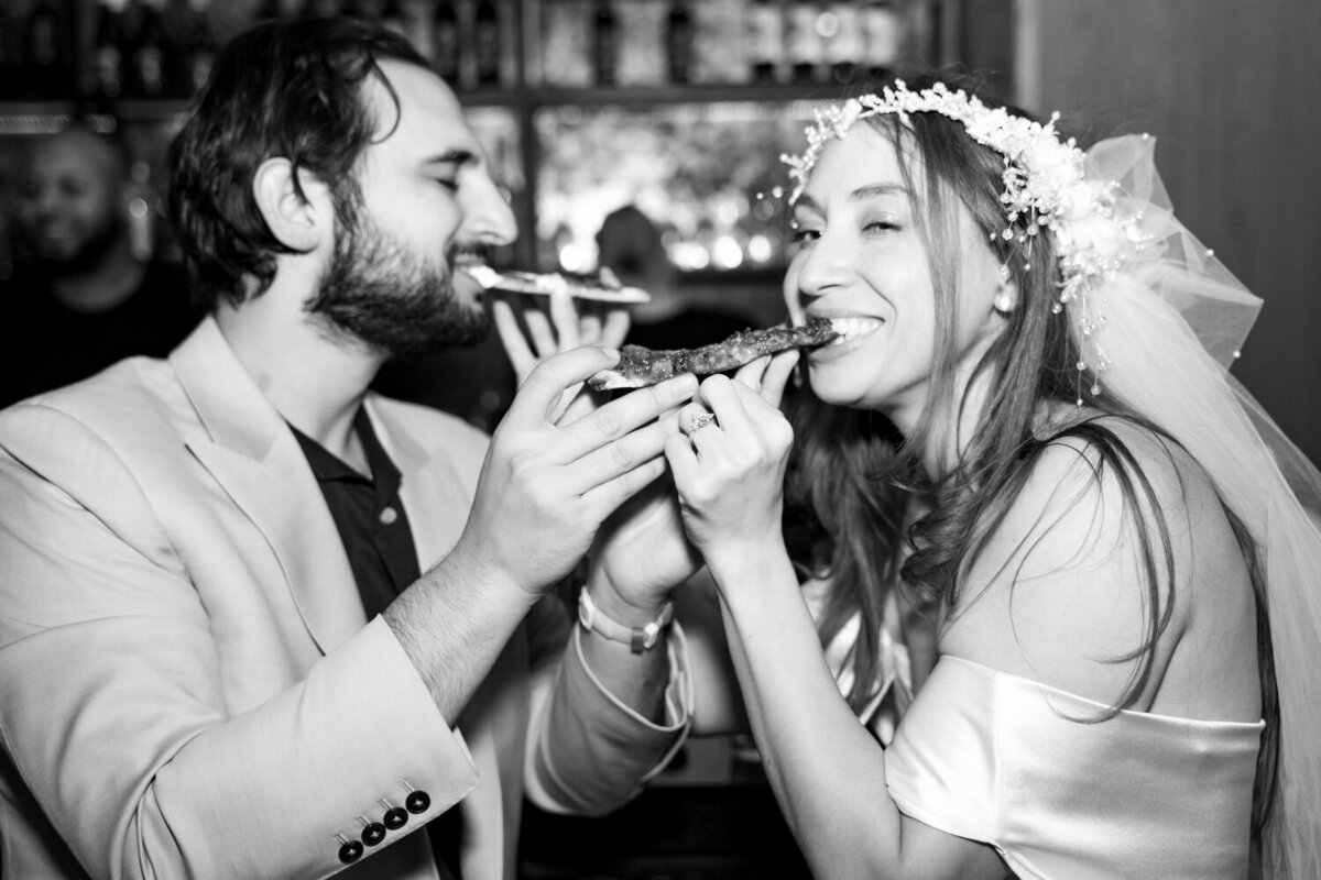 paparazzi-style black and white photo of a couple feeding each other pizza during a rehearsal dinner