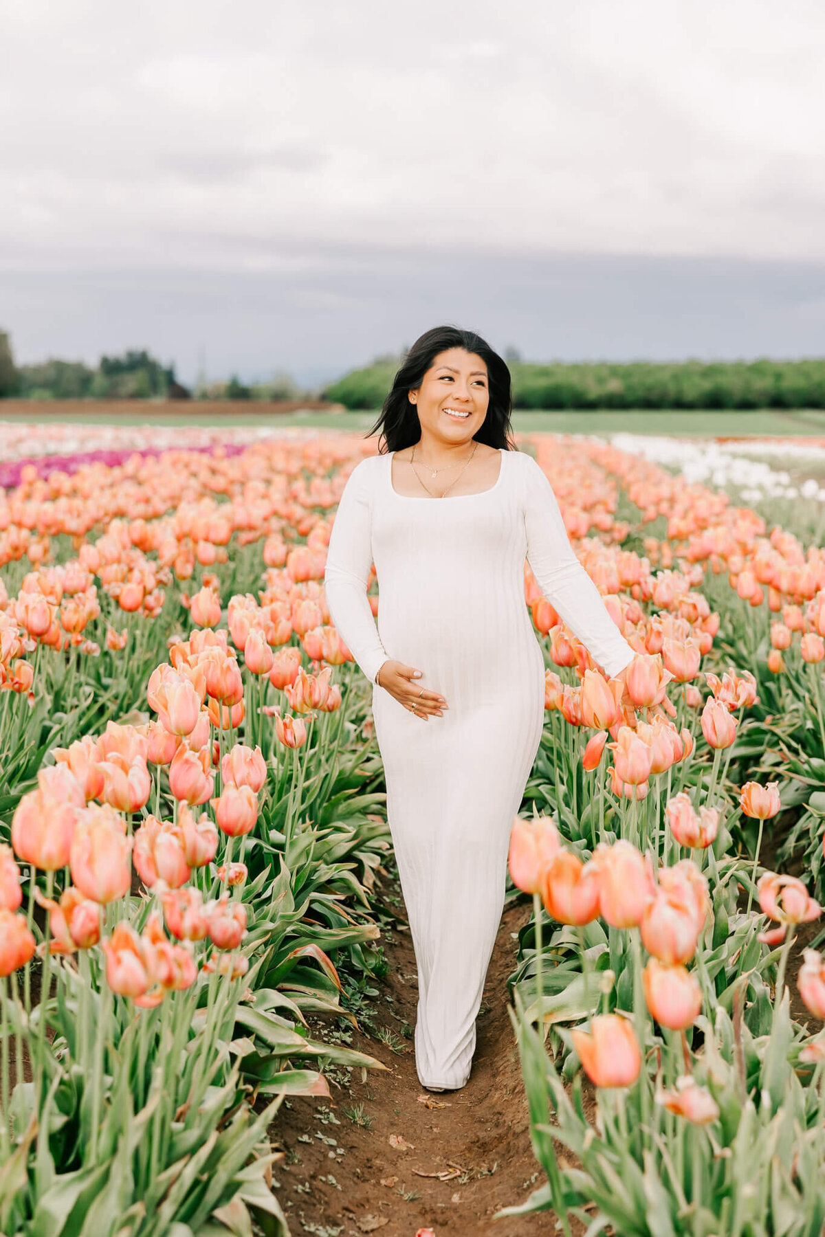 Joyful woman walking and smiling in the tulip fields in Woodburn oregon.  There are pink tulips on both sides of her and she has black hair and a white dress on.