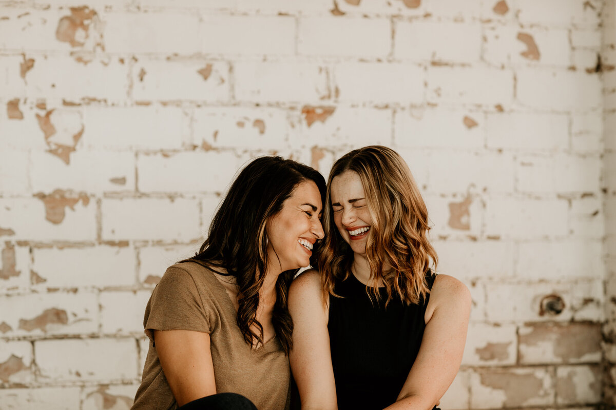 same sex couple laughing  and sitting together in front of a white brick wall