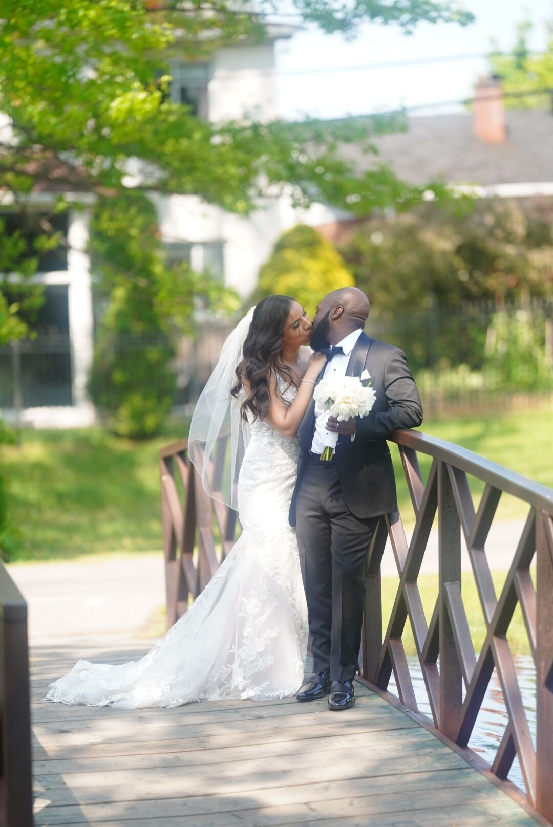 A couple shares a tender kiss on a scenic bridge in a nature park, surrounded by lush greenery and tranquil waters. The image captures a romantic moment amid the natural beauty of the park, reflecting the couple's love and connection.