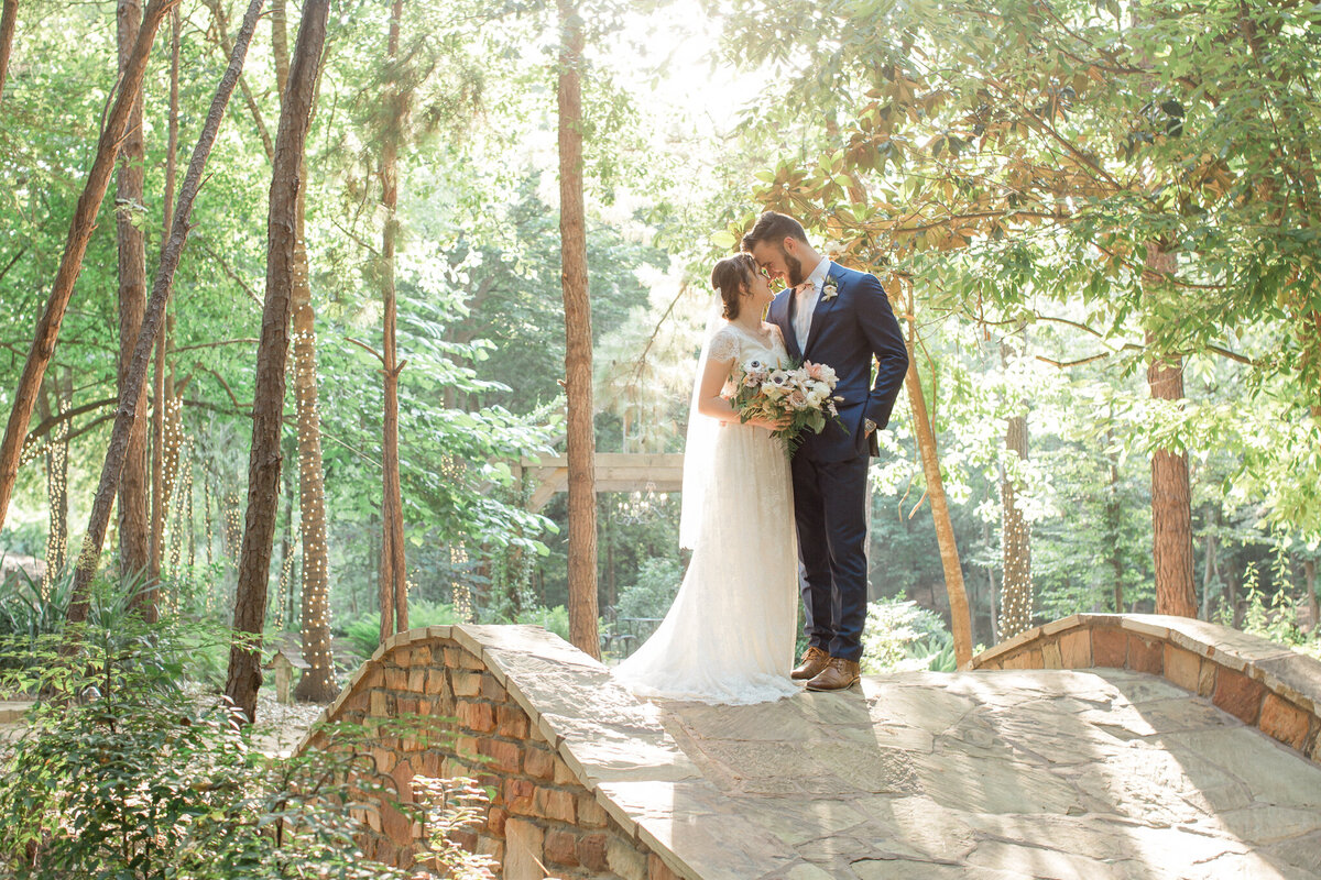 The bride and groom stand on a stone bridge in the woods, noses touching and looking into each others eyes