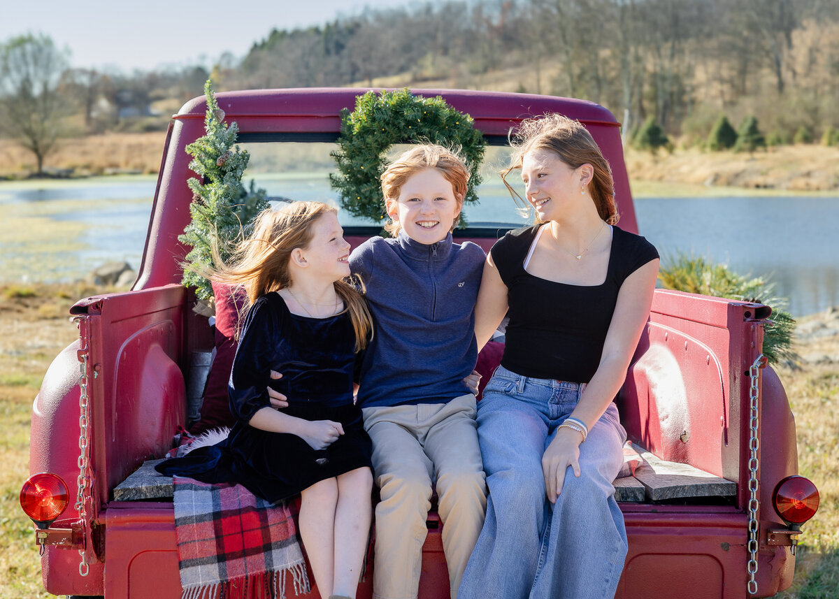 Sibling photo on a red pickup truck decorated for the holidays at a Christmas tree farm in Somerset County, NJ