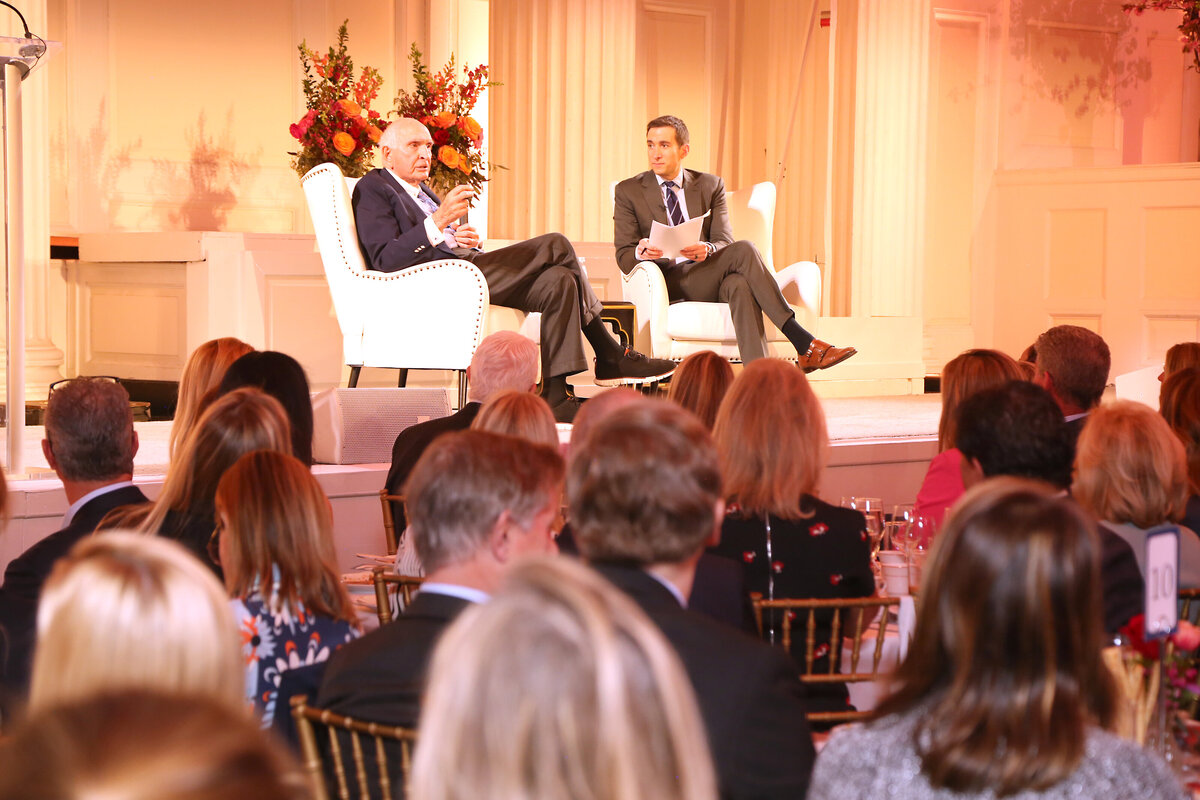 Speaker and host seated on white armchairs having a conversation on stage before an audience during the BCNY Annual Luncheon 2025 in New York.
