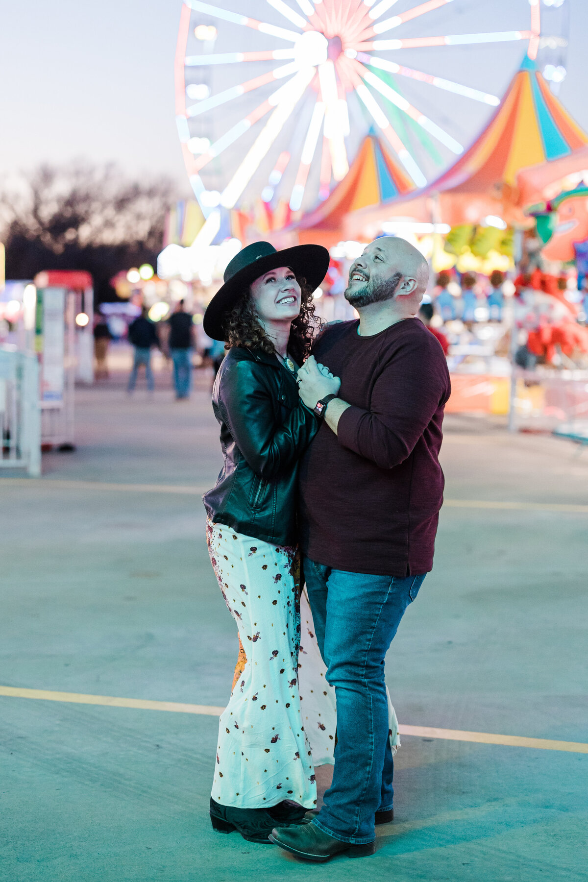 A couple dancing and smiling together at a carnival during their engagement photoshoot in DFW, Texas. The woman on the left is wearing a black brimmed hat, a black leather jacket, and a detailed, long white skirt. The man on the right is wearing a maroon sweater and jeans. A colorful, lively carnival setting can be seen behind them including a large ferris wheel.