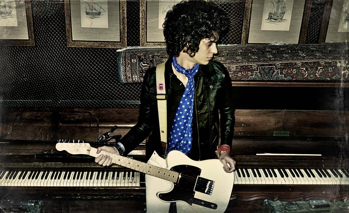 Male musician photo wearing black leather jacket white pants holding white electric guitar sitting against old wood piano