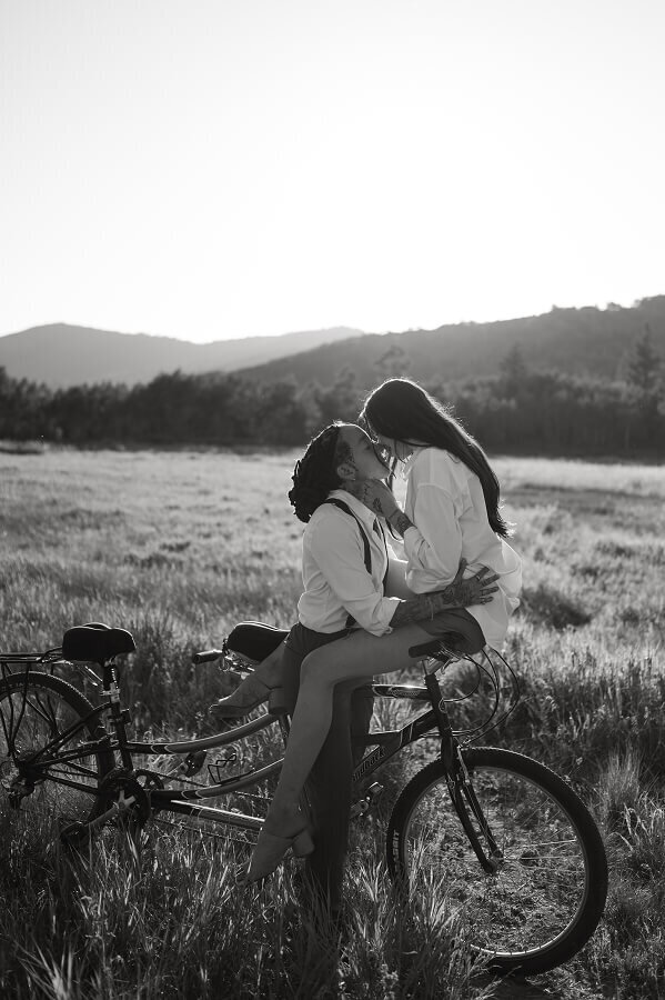A timeless black and white engagements photo of a woman kissing her fiancé. She is balancing on the handlebars of a bicycle. 