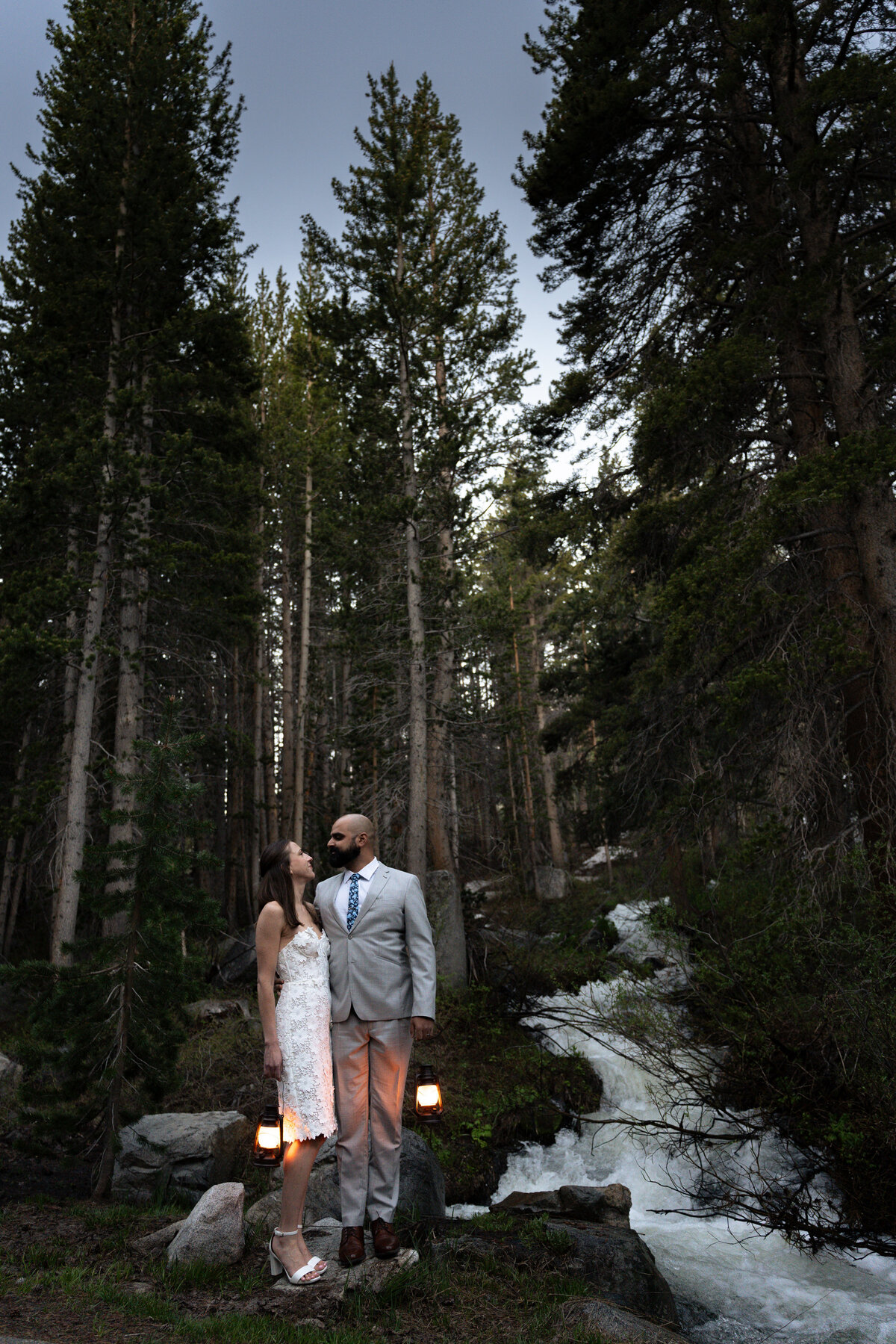 A couple holding lanterns and standing next to a rushing river in the forest