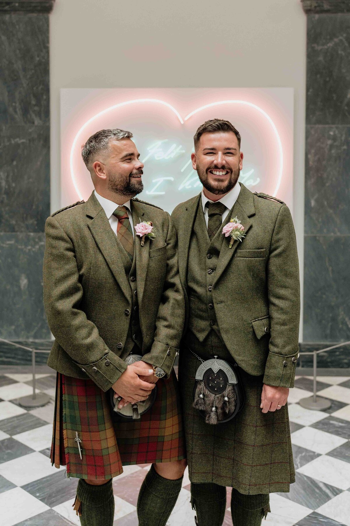 Two grooms are photographed smiling and wearing traditional Scottish kilts and suits in Aberdeen Art Gallery. They stand on a checkered floor with a neon heart-shaped sign in the background, both adorned with boutonnieres on their jackets.