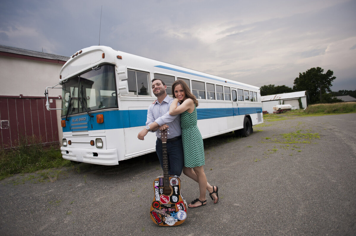 A couple leaning on each other in front of a bus while one holds a guitar 