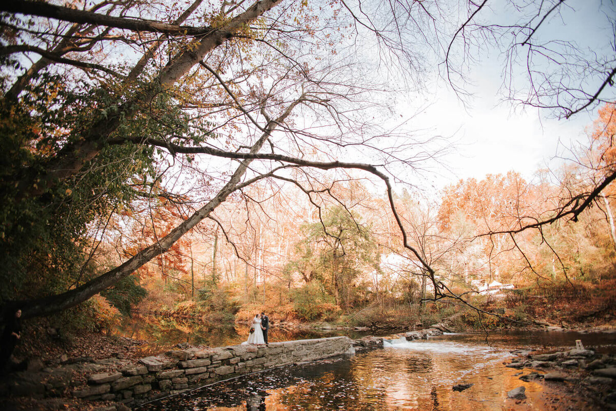 A couple stands on a stone path beside a reflective stream, surrounded by vibrant autumn trees at Valley Green Inn, Philadelphia.