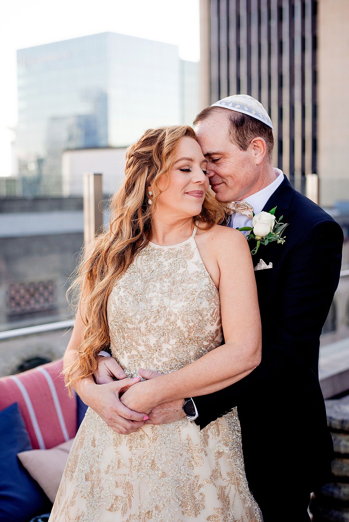Jewish groom wearing a blakc tuxedo and white kippot and Jewish Bride wearing a gold halter top beaded wedding dress embrace on a Nashville Rooftop at Noelle Hotel at their Jewish Wedding