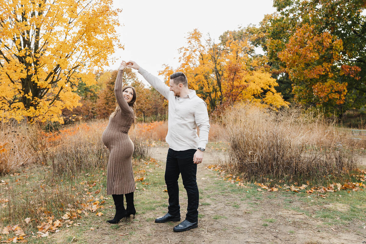 Husband twirling his pregnant wife during fall maternity session in High Park. Mom is dressed in fitted knit brown dress with tall black boots.